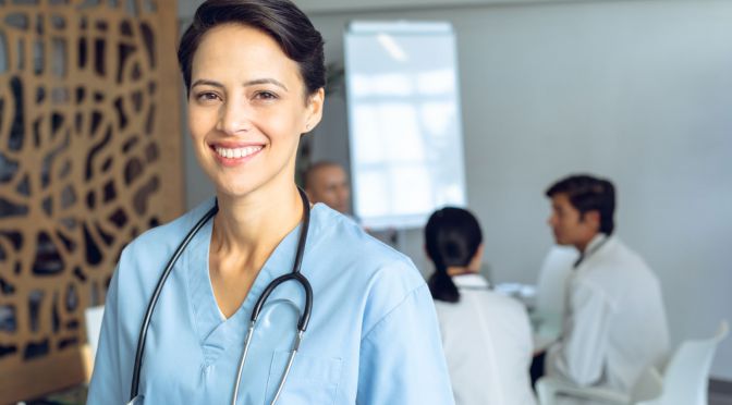 Front view of happy female Caucasian nurse looking at camera in conference room in hospital. Diverse colleagues in meeting sitting at the table of conference room in background. Front view of happy female Caucasian nurse looking at camera in conference room in hospital. Diverse colleagues in meeting sitting at the table of conference room in background.