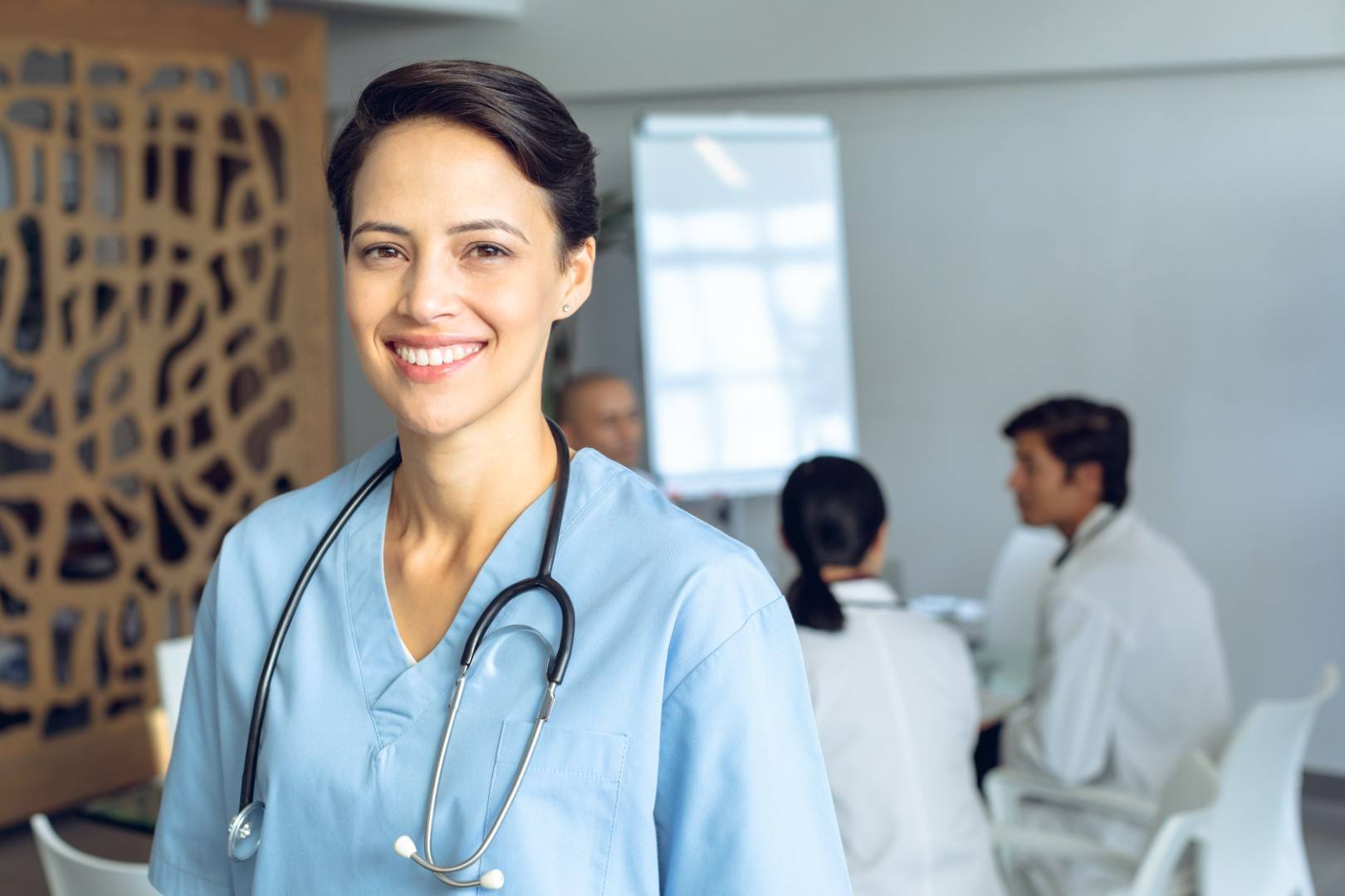 Front view of happy female Caucasian nurse looking at camera in conference room in hospital. Diverse colleagues in meeting sitting at the table of conference room in background.